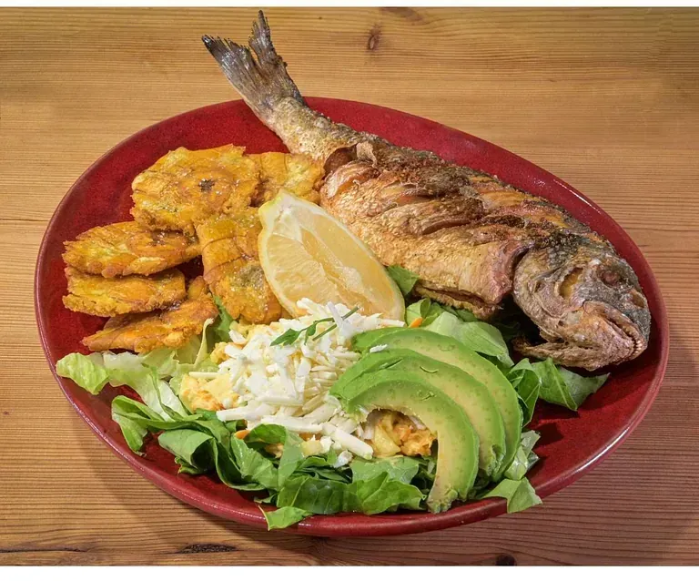 Un plato rojo cubierto con pescado frito, aguacate y plátanos sobre una mesa de madera.
