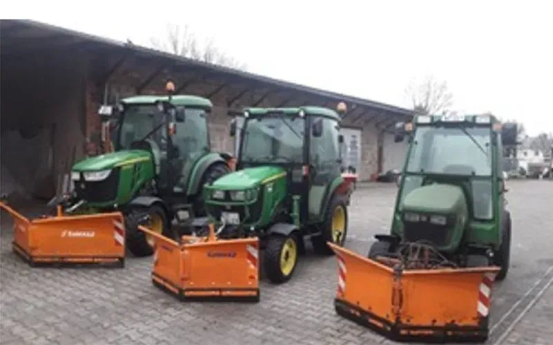 Drei John Deere-Traktoren mit Schneepflügen sind vor einem Gebäude geparkt.