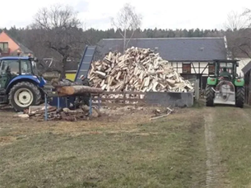 Zwei Traktoren schneiden Holz auf einem Feld vor einem Haus.