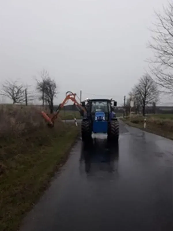 Ein blauer Traktor fährt eine nasse Straße entlang. Gala Bau & Service GbR