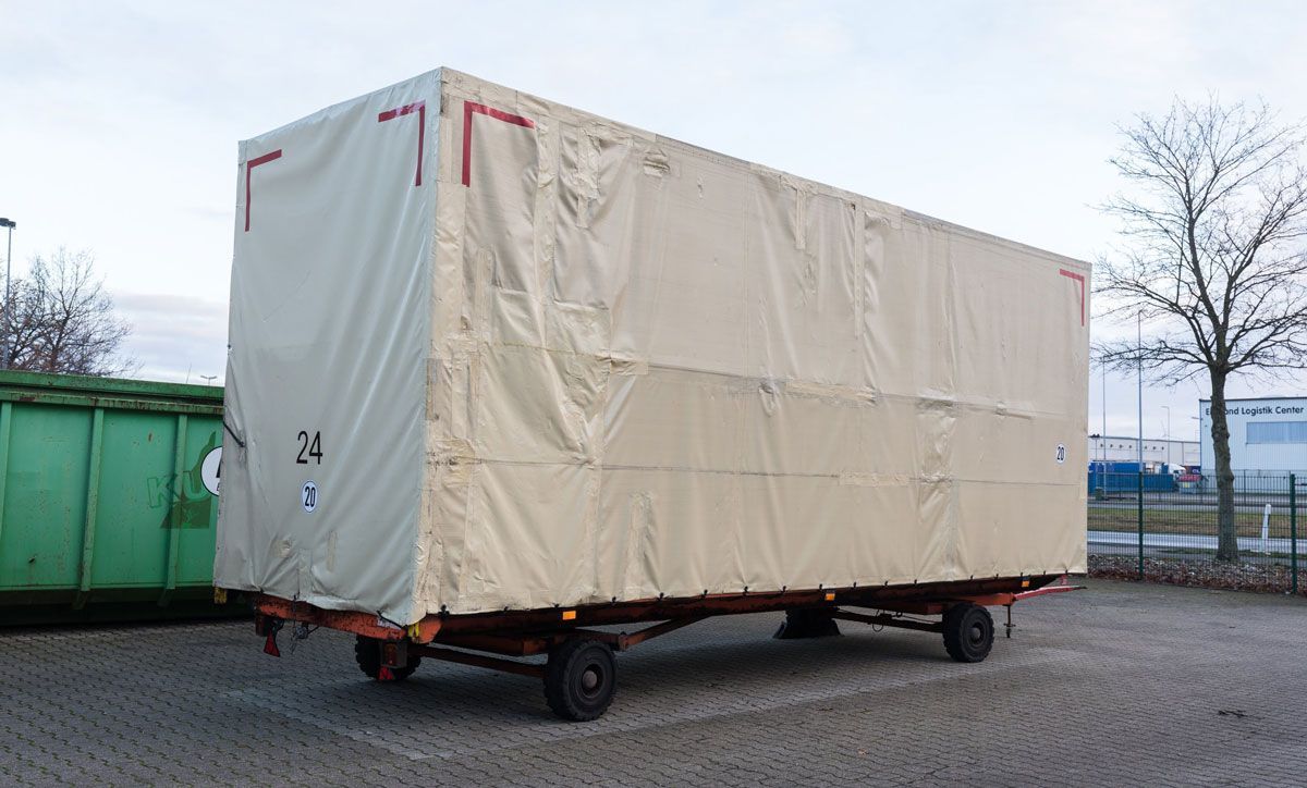 Ein großer weißer Anhänger steht auf einem Parkplatz neben einem grünen Müllcontainer.