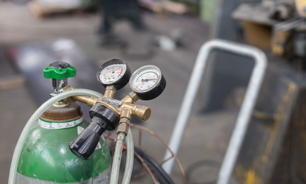 Auf einem Wagen steht eine grüne Gasflasche mit zwei daran befestigten Messgeräten.