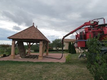Pavillon en briques à côté d'une machine agricole rouge pulvérisant un liquide sur un champ herbeux sous un ciel nuageux.