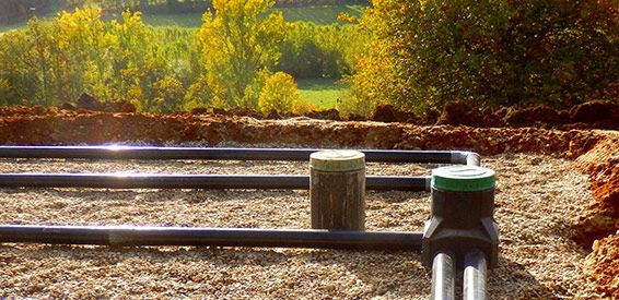 Des barres de clôture en bois avec des poteaux aux chapeaux verts bordent un chemin de terre ensoleillé et des arbres d'automne.