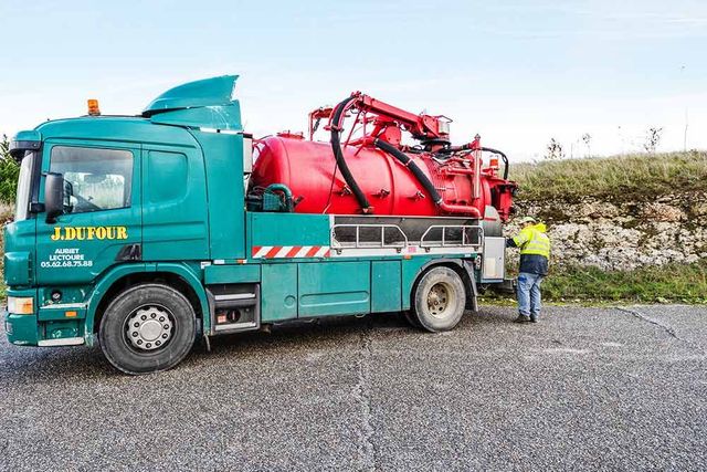 Un camion de dépannage turquoise transportant un réservoir rouge est stationné au bord d'une route de gravier, un ouvrier se tenant à côté.