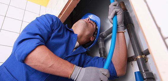 Un technicien en uniforme bleu inspecte des canalisations à l'aide d'une lampe torche dans une zone technique carrelée.