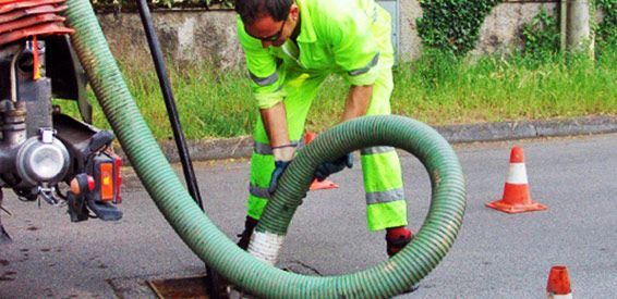 Un ouvrier en tenue vert fluo manipule des tuyaux sur un site de nettoyage routier délimité par des cônes de signalisation orange.