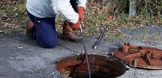 Un ouvrier agenouillé près d'une bouche d'égout ouverte utilise un outil sur un câble en bordure de la chaussée.