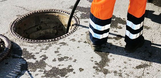 Un ouvrier en pantalon de sécurité orange se tient près d'une bouche d'égout ouverte sur une surface en béton.