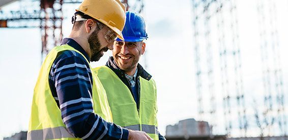 Deux ouvriers du bâtiment, casqués et vêtus de gilets de sécurité, examinent des plans sur un chantier.