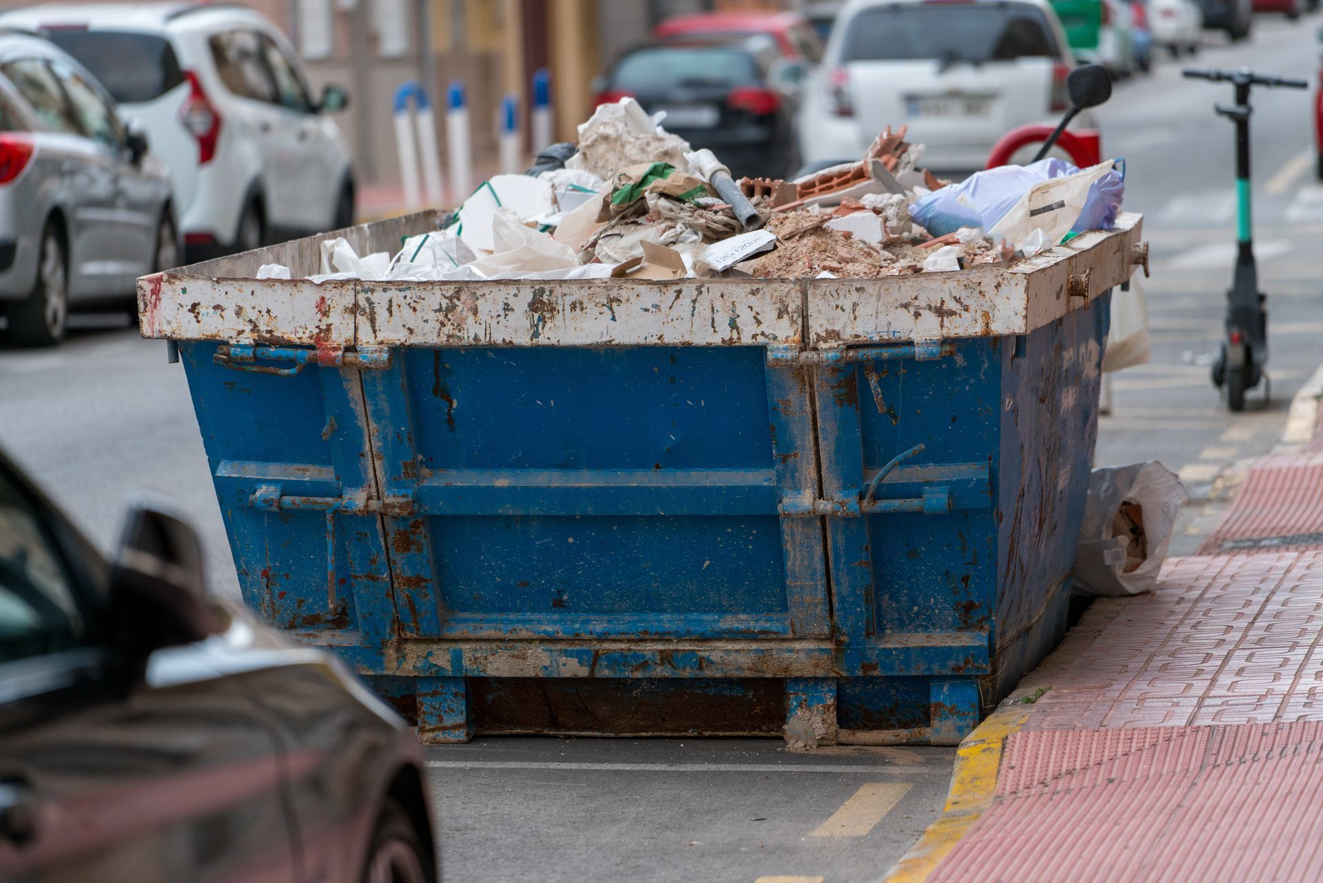 Une benne à ordures bleue débordant de déchets se trouve dans une rue de la ville, à côté de voitures stationnées.
