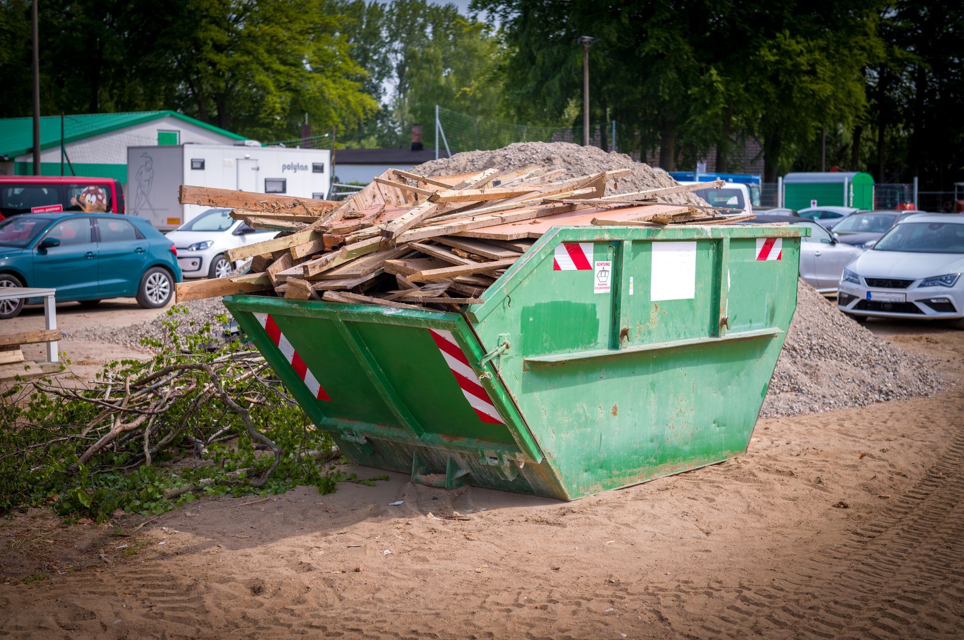 Une benne verte remplie de débris de bois, avec des voitures et des arbres en arrière-plan.