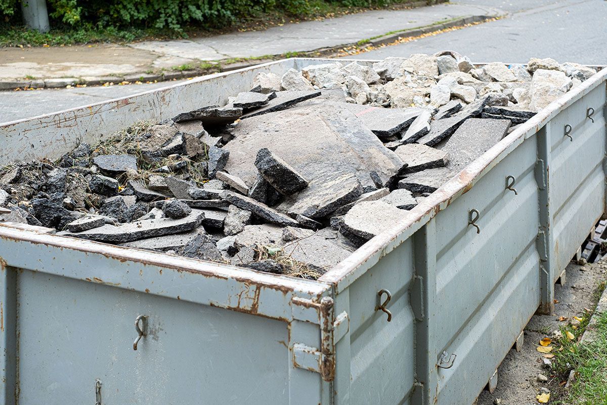 Un conteneur gris rempli de gravats de béton cassés, en bordure de trottoir.