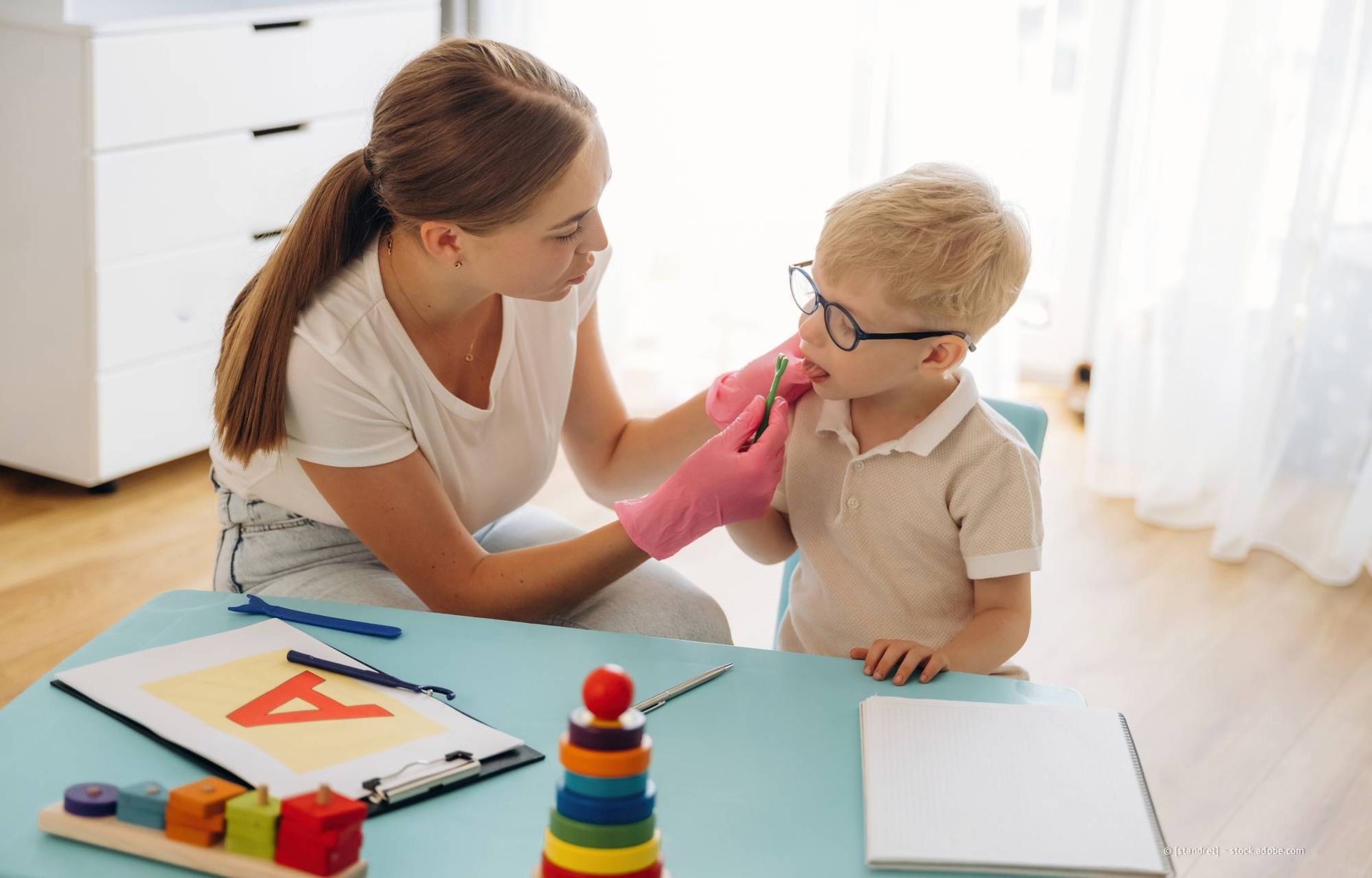 Eine Frau mit behandschuhter Hand untersucht den Mund eines Kindes an einem Tisch mit Lernmaterialien.