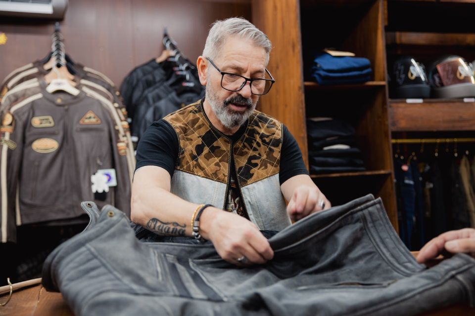 Ein Lederhandwerker begutachtet eine schwarze Jacke in einem Geschäft, hinter dem Jacken hängen.