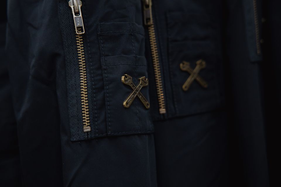 Nahaufnahme einer dunkelblauen Jacke mit goldenen Reissverschlüssen und gekreuzten Lederriemen auf der Vorderseite.
