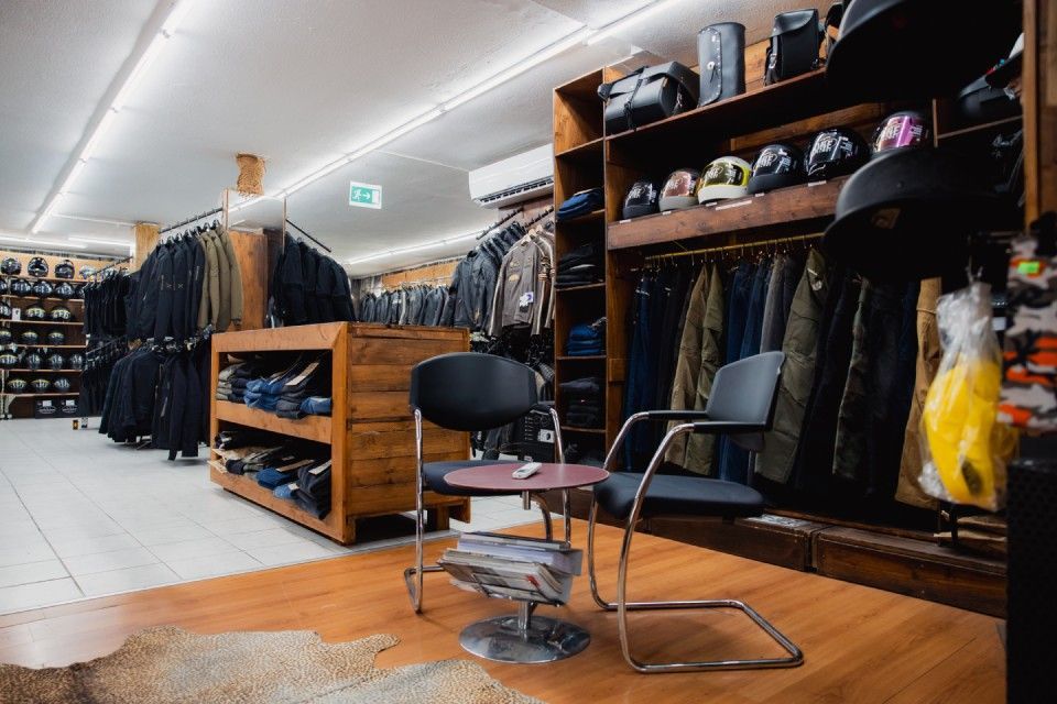 Innenansicht eines Bekleidungsgeschäfts mit Jackenständern, Schuhregalen und zwei Stühlen auf einem Holzboden.