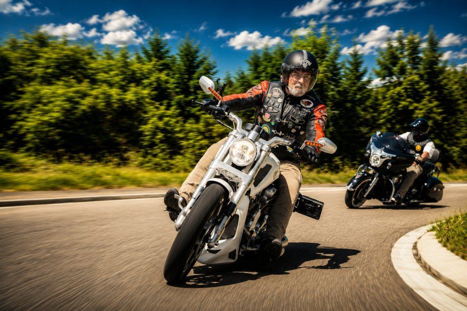 Zwei Motorräder neigen sich durch eine sonnige Kurve auf einer von Bäumen gesäumten Strasse.