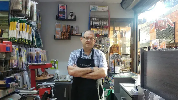 Persona de pie con los brazos cruzados en una pequeña tienda, rodeada de mercancía colgada y estantes.