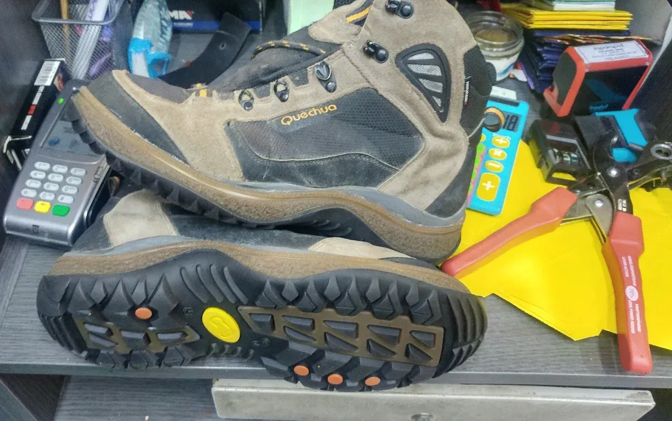 Un par de botas de montaña desgastadas sobre un banco de trabajo desordenado; una de ellas muestra la suela y la parte superior de color marrón grisáceo.