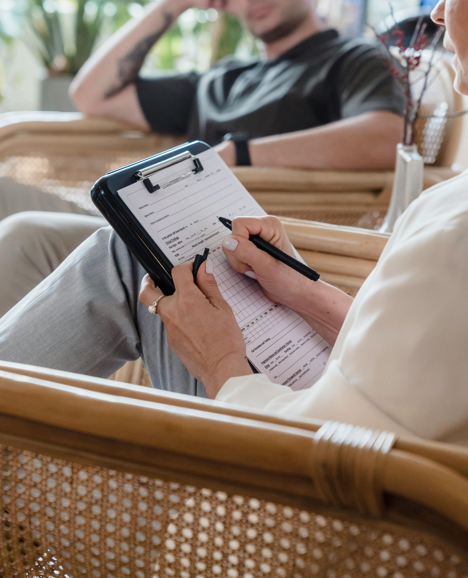 Una persona escribe en un portapapeles sentada en una silla de mimbre, con otra sentada al fondo.