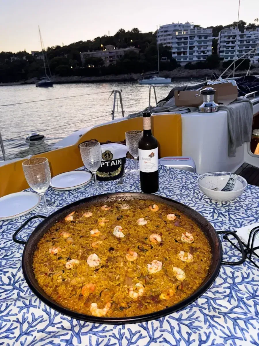 Paella servida en una mesa de barco al atardecer, con vino y edificios del paseo marítimo de fondo.