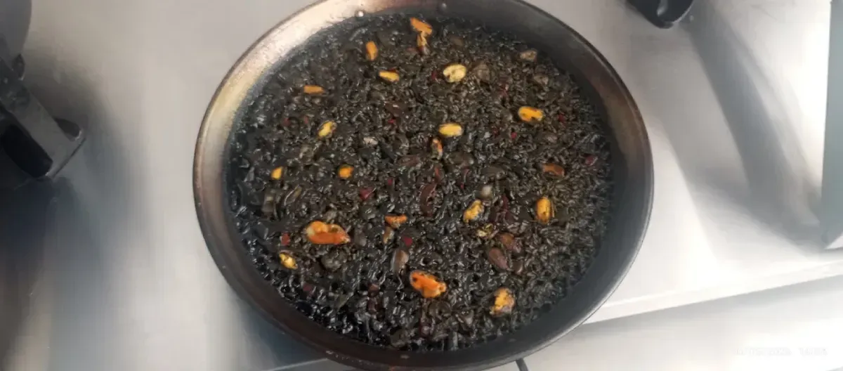 Sartén negra llena de material granular oscuro y pequeños trozos naranjas sobre una superficie blanca.