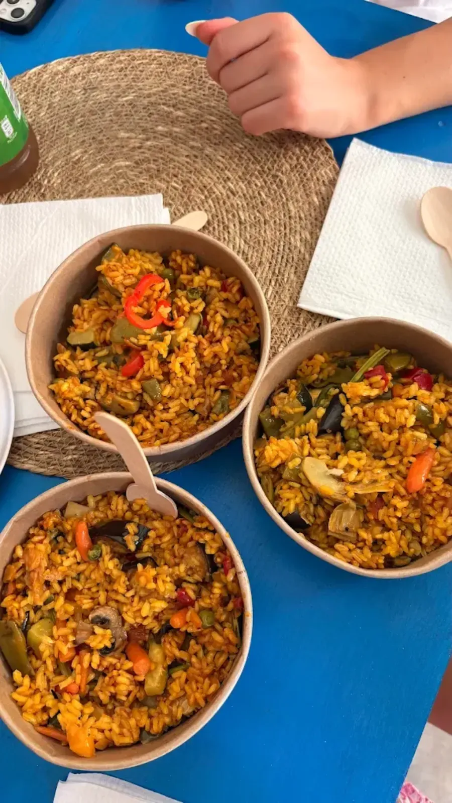 Tres cuencos de arroz y verduras sobre una mesa azul, con la mano de un niño cerca.