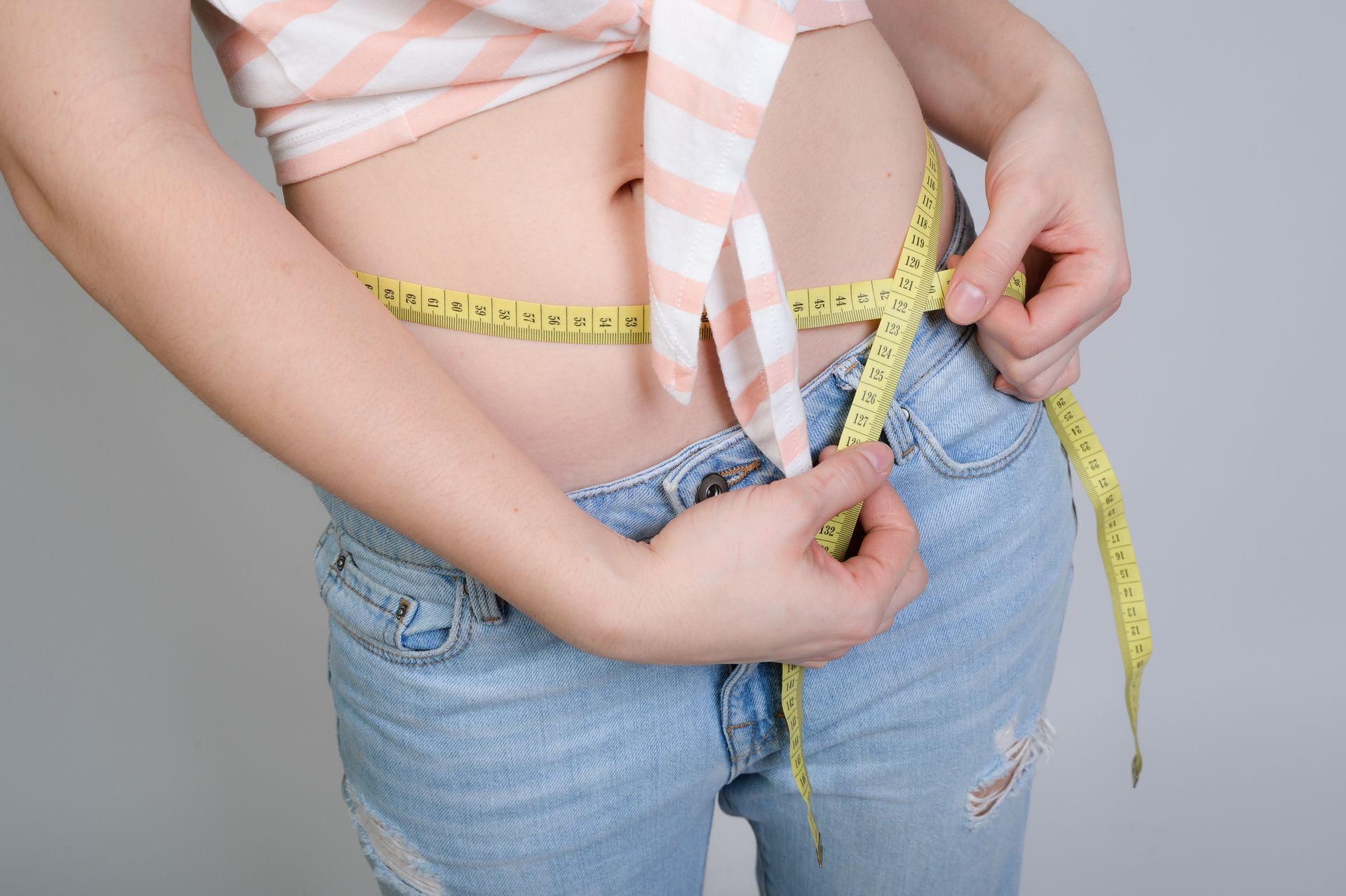 Une personne mesure son tour de taille avec un mètre ruban jaune, vêtue d'un jean et d'un haut court.