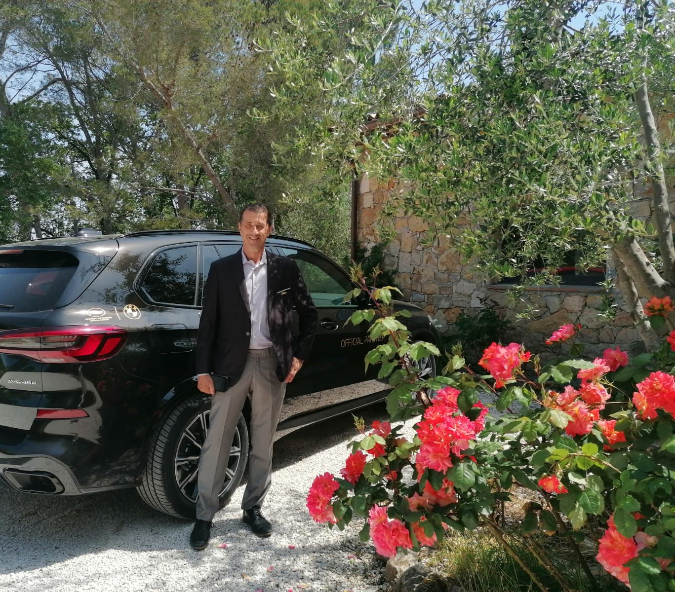 Un homme se tient debout à côté d'un SUV noir dans une allée ensoleillée, avec des fleurs rouges au premier plan.