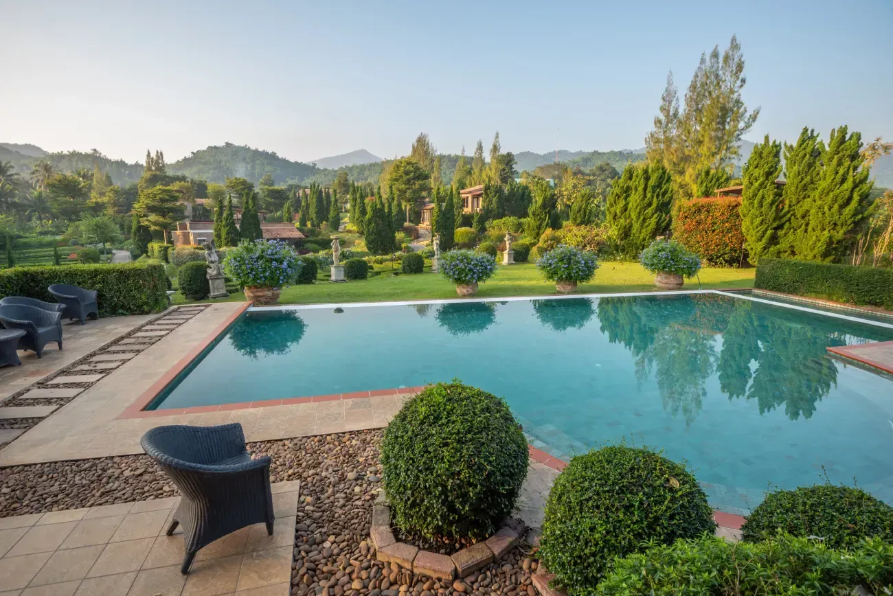 Una piscina rodeada de un jardín impecablemente cuidado, con arbustos esféricos, árboles y montañas al fondo.