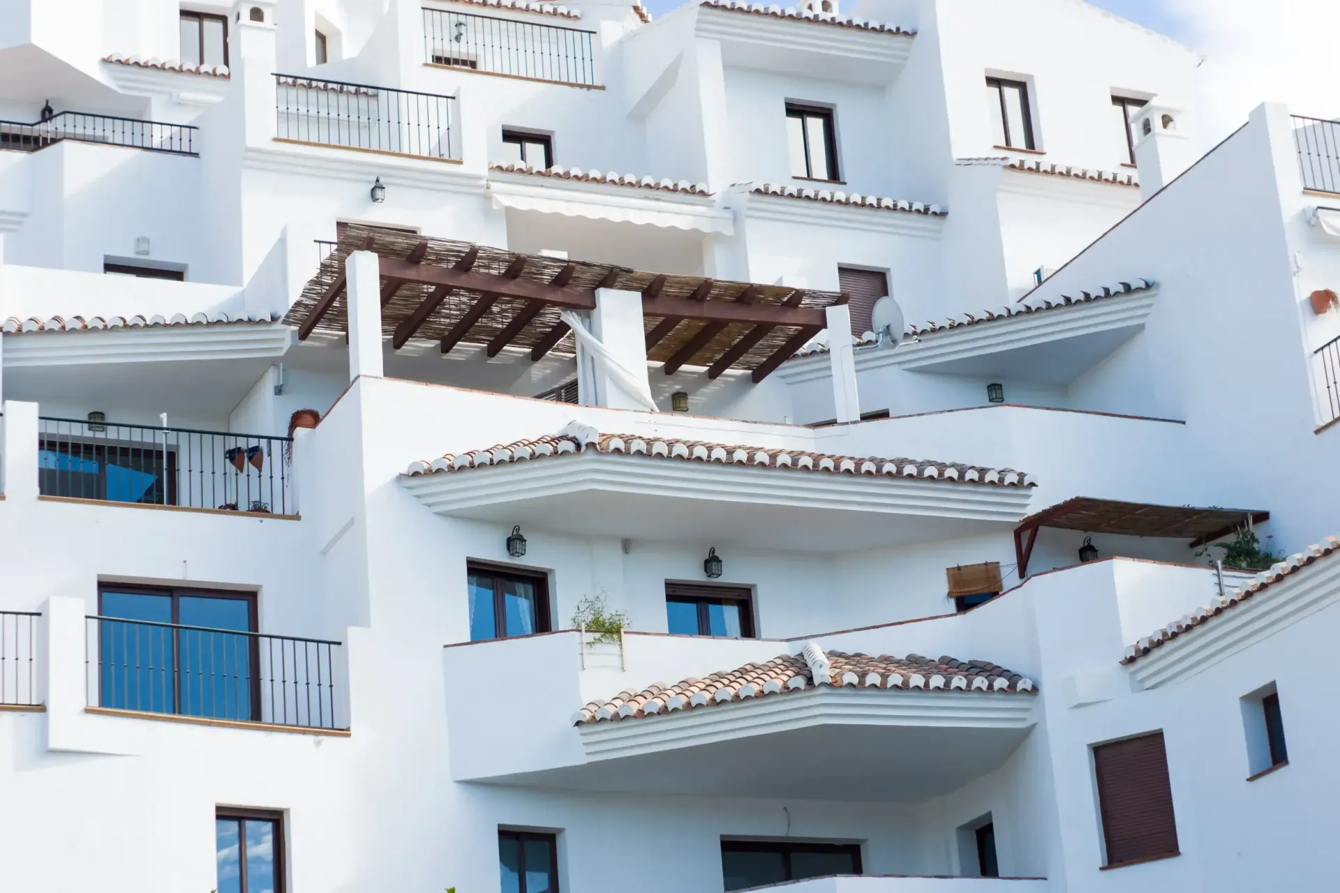 Un edificio blanco de varias plantas de estilo mediterráneo, con balcones escalonados, ventanas oscuras y pérgolas de madera.