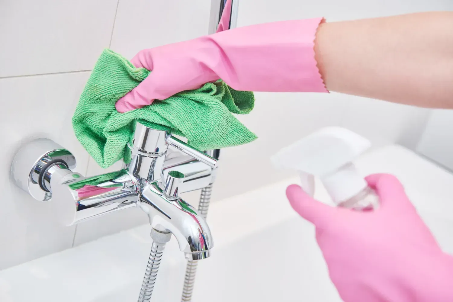 Manos con guantes de goma rosas limpiando un grifo de baño cromado con un paño verde y una botella pulverizadora.