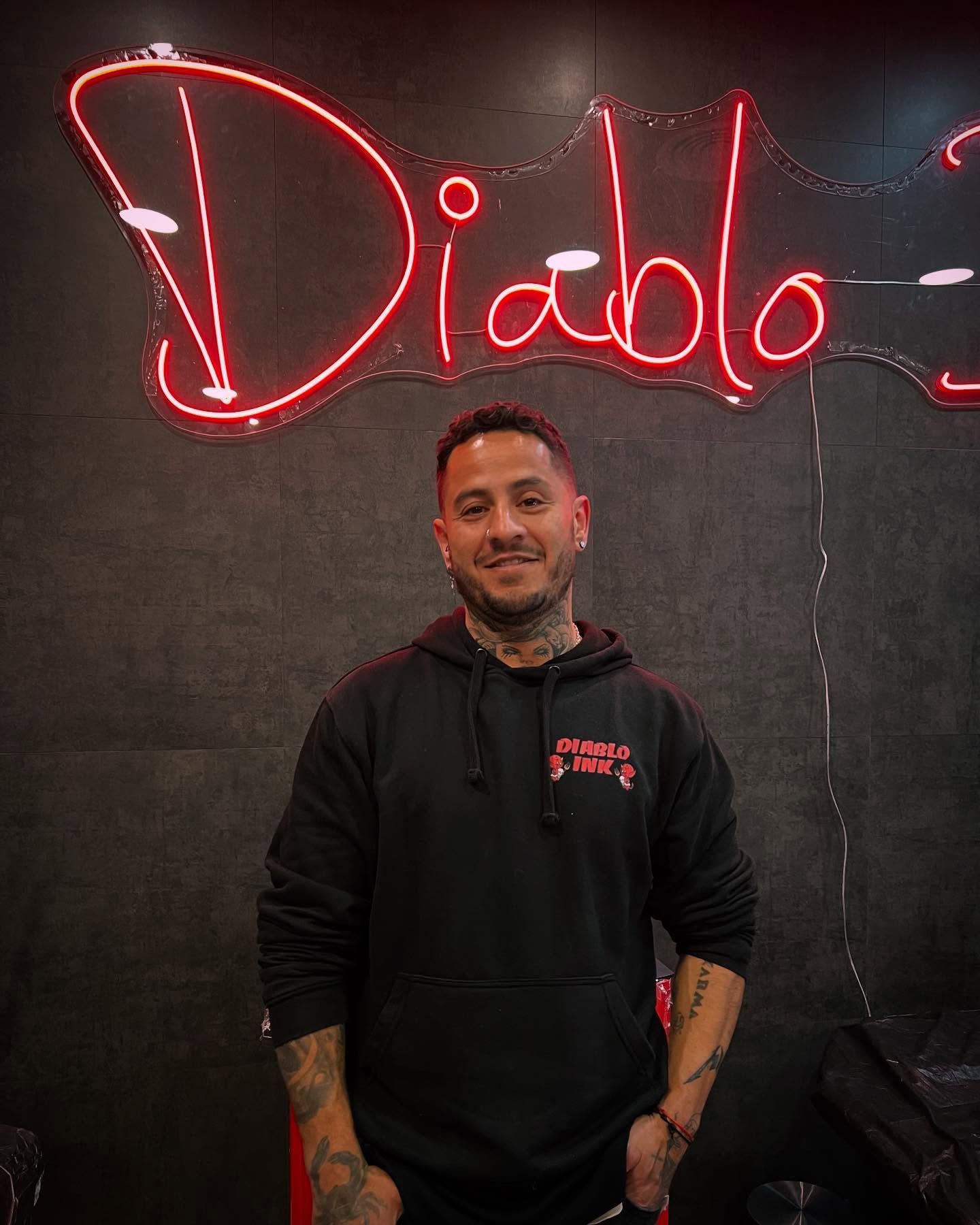 Un hombre con tatuajes y sudadera con capucha negra sonríe frente a un cartel de neón que dice