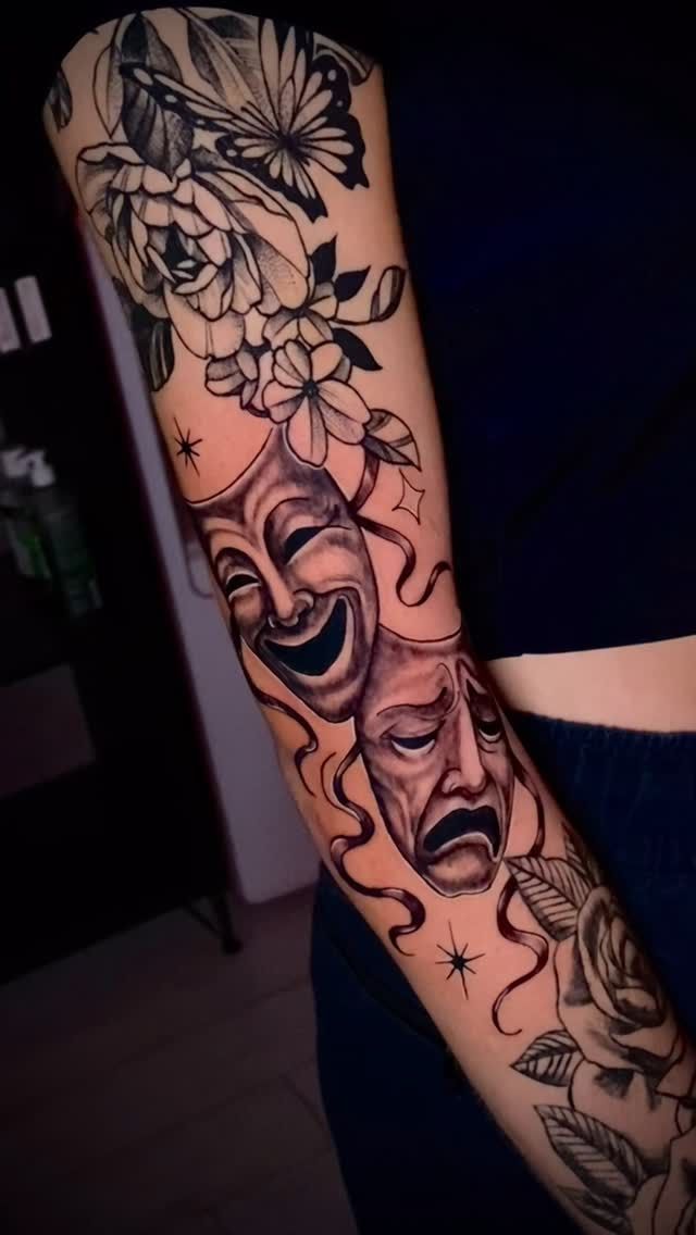 Tatuaje en el brazo: máscaras de comedia y tragedia con flores; tinta negra sobre piel.