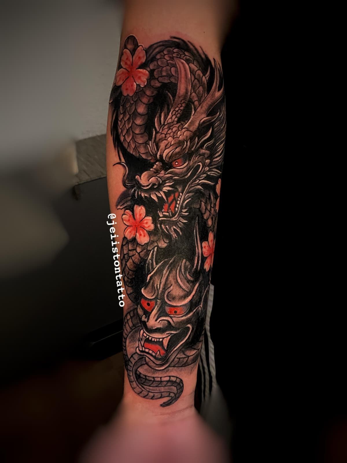 Tatuaje de máscara de dragón y demonio con acentos rojos y flores de cerezo en el brazo.