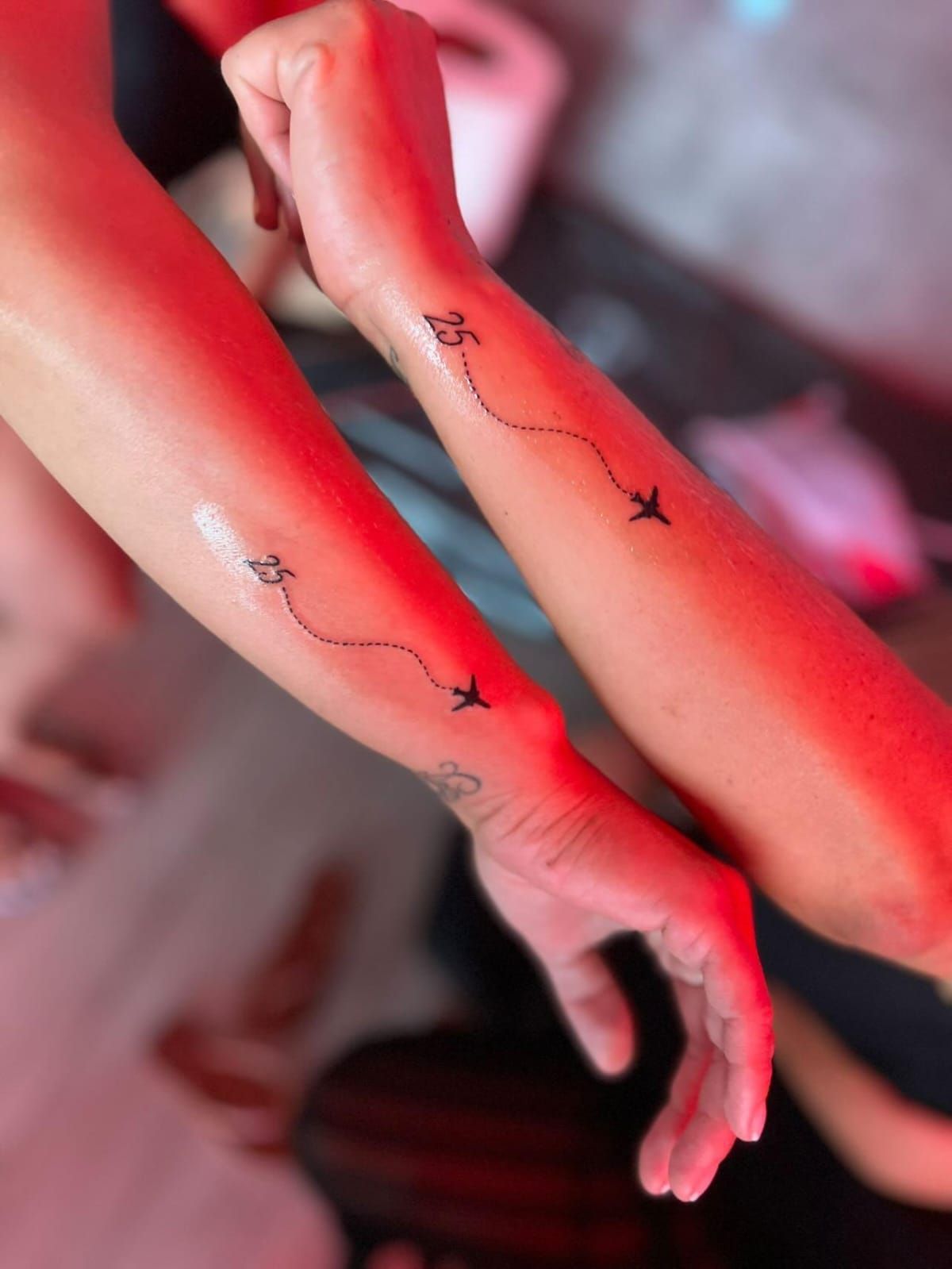 Dos brazos con tatuajes de aviones a juego conectados por líneas de puntos, iluminación roja.