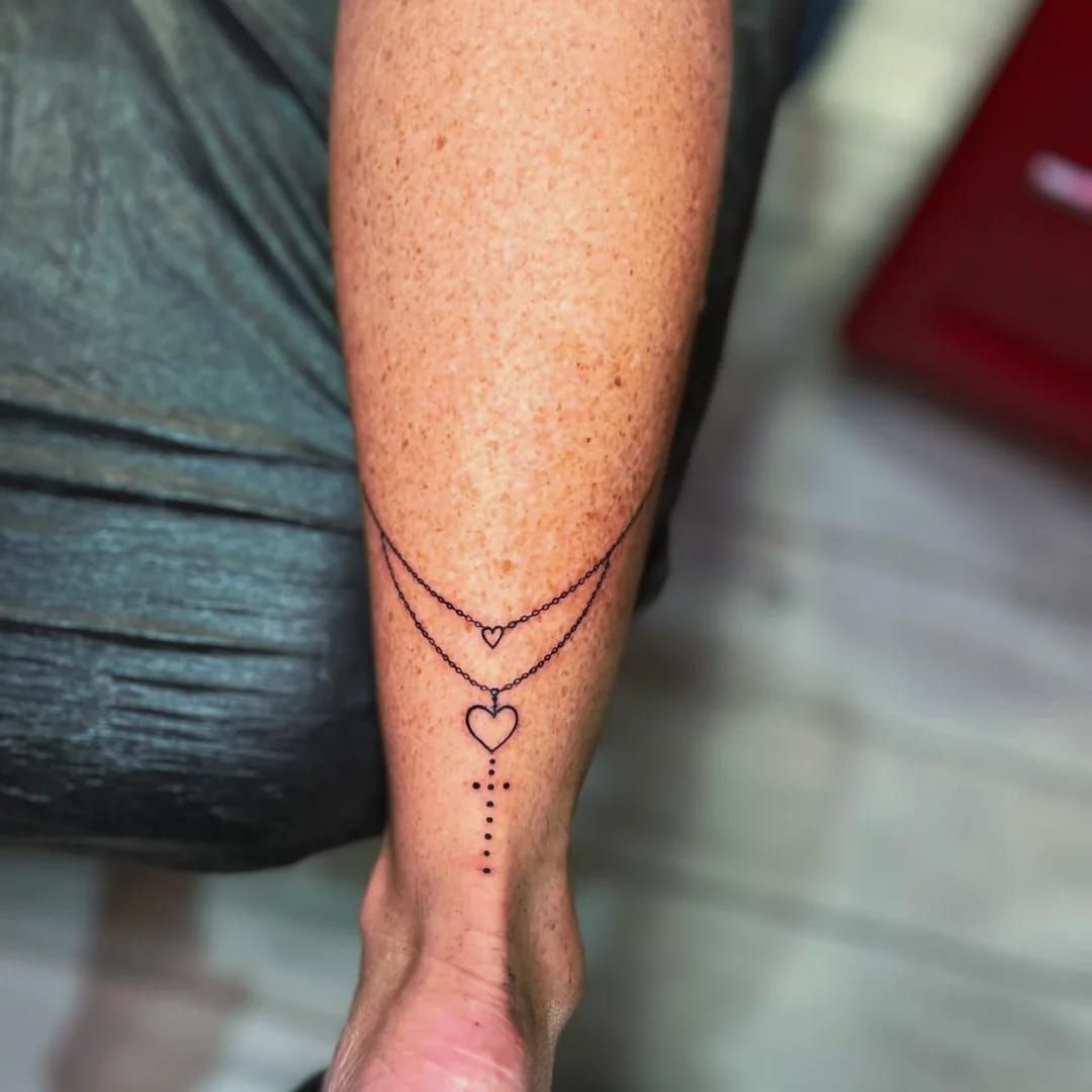 Tatuaje de un diseño de collar en un antebrazo con un corazón, una cruz y un corazón pequeño.