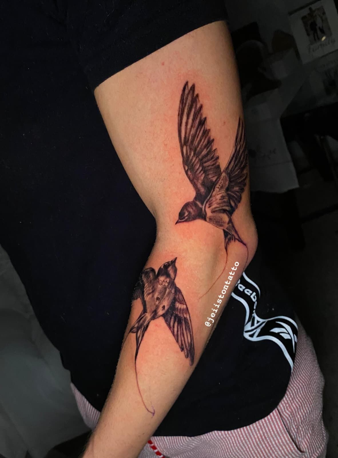 Dos tatuajes de pájaros negros en el brazo de una persona, uno encima del codo y otro debajo, en vuelo.