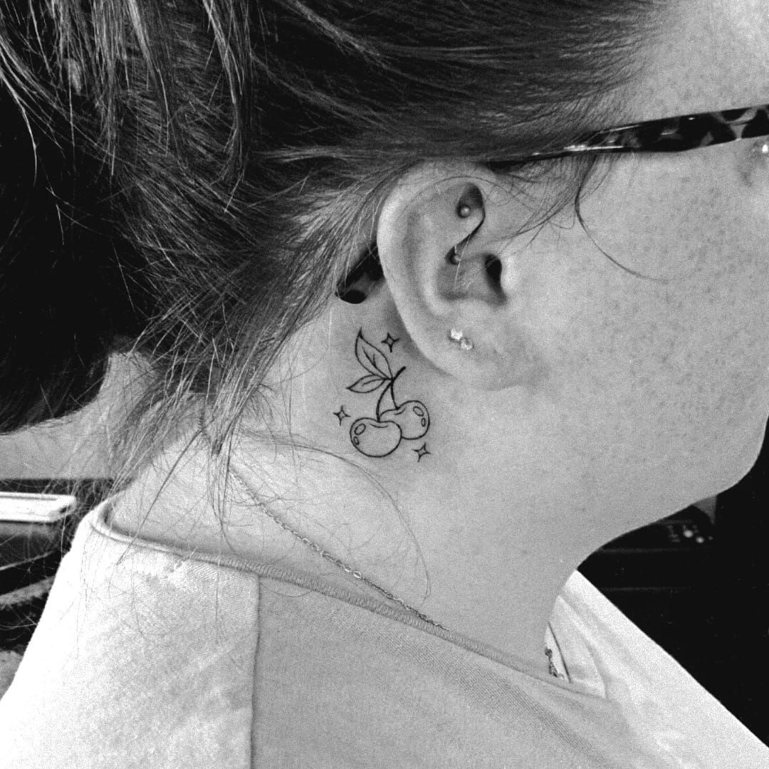 Fotografía en blanco y negro de un tatuaje en el cuello de unas cerezas con algunas estrellas, junto a la oreja de una persona con gafas.