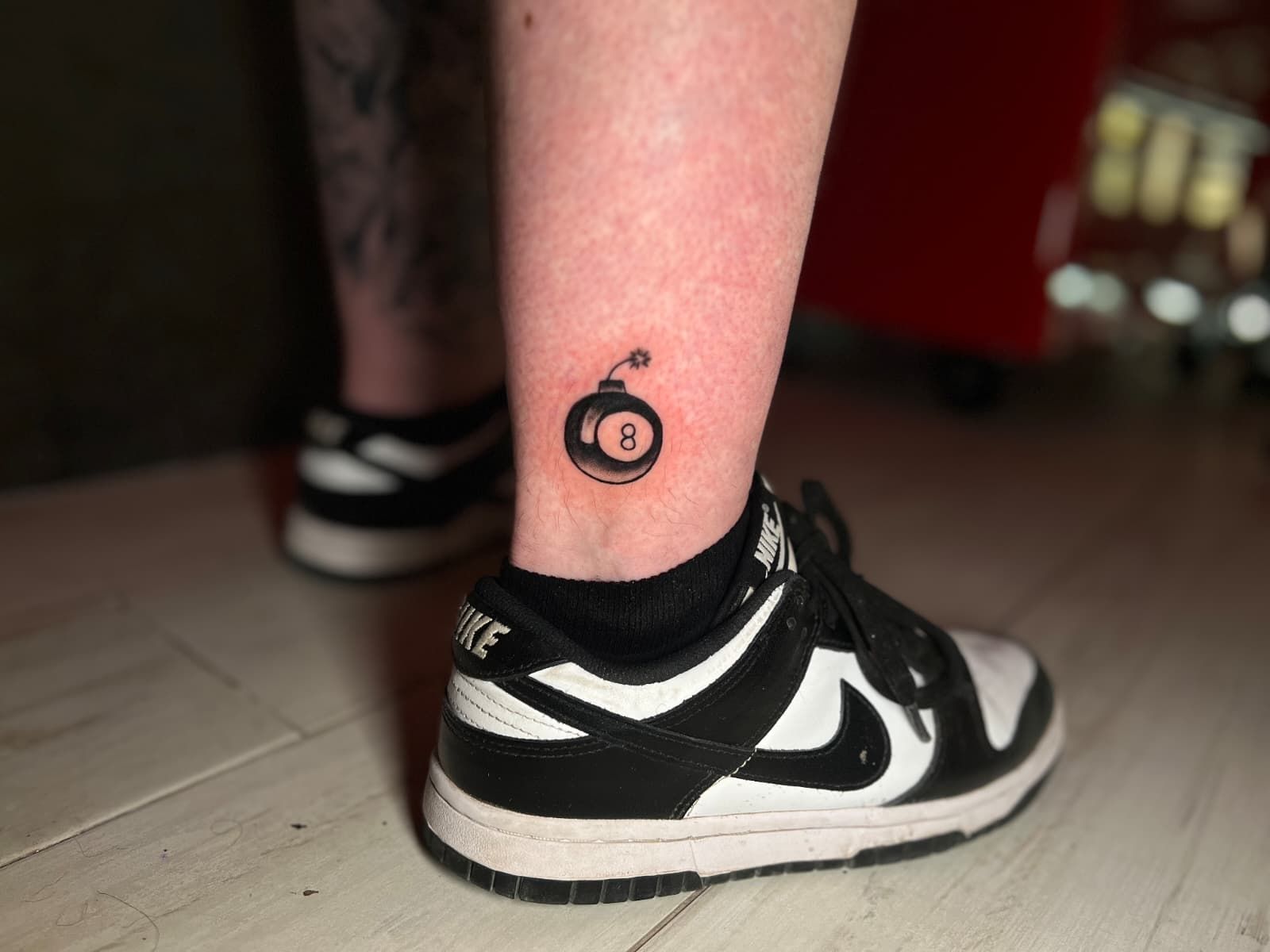 Tatuaje en el tobillo de una bola 8 como bomba, encima de una zapatilla Nike Dunk en blanco y negro.