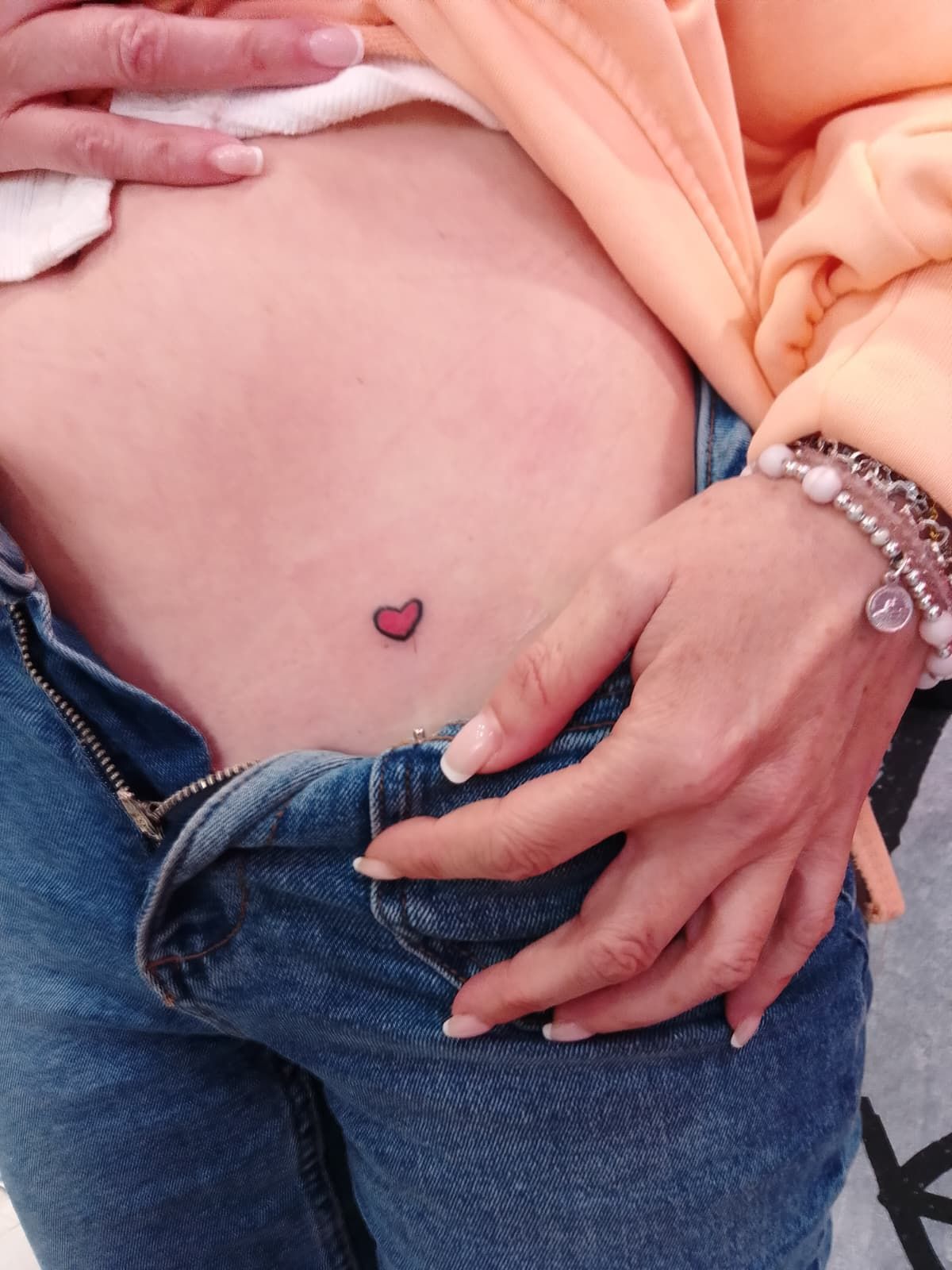Cintura de una persona con un pequeño tatuaje de corazón rojo, jeans desabrochados y una pulsera rosa.