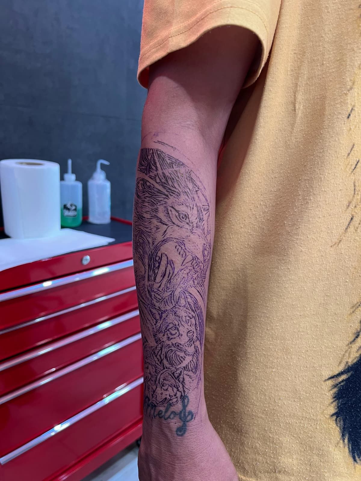 Brazo de una persona con un tatuaje de diseño floral/orgánico detallado. Lleva una camisa amarilla, cerca de una caja de herramientas roja y suministros.