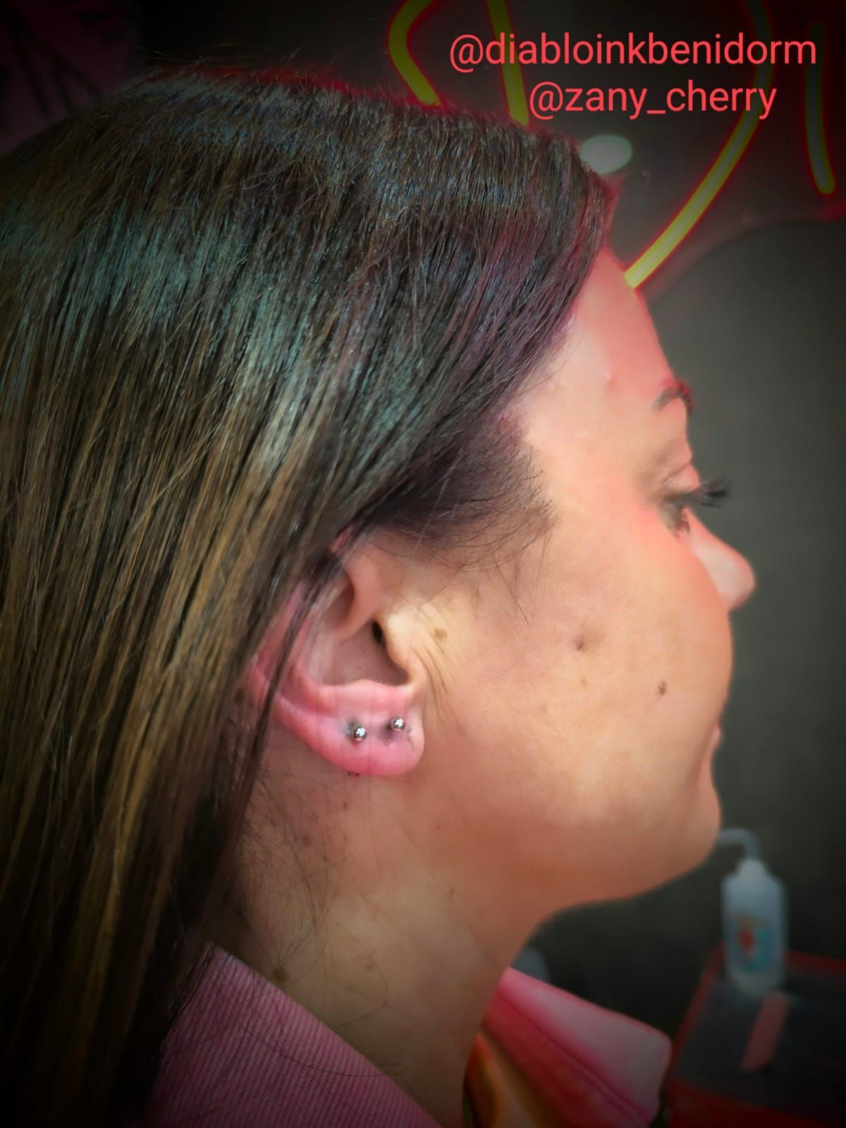 Mujer de perfil; se ven dos piercings en las orejas. Vestida con ropa rosa, en un entorno con poca luz.