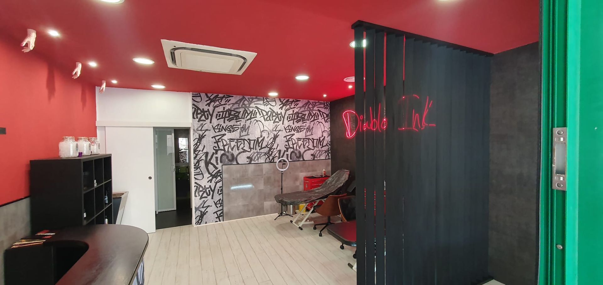 Interior de un salón de tatuajes: techo rojo, arte mural en blanco y negro, silla de tatuajes, estanterías negras y un biombo negro.