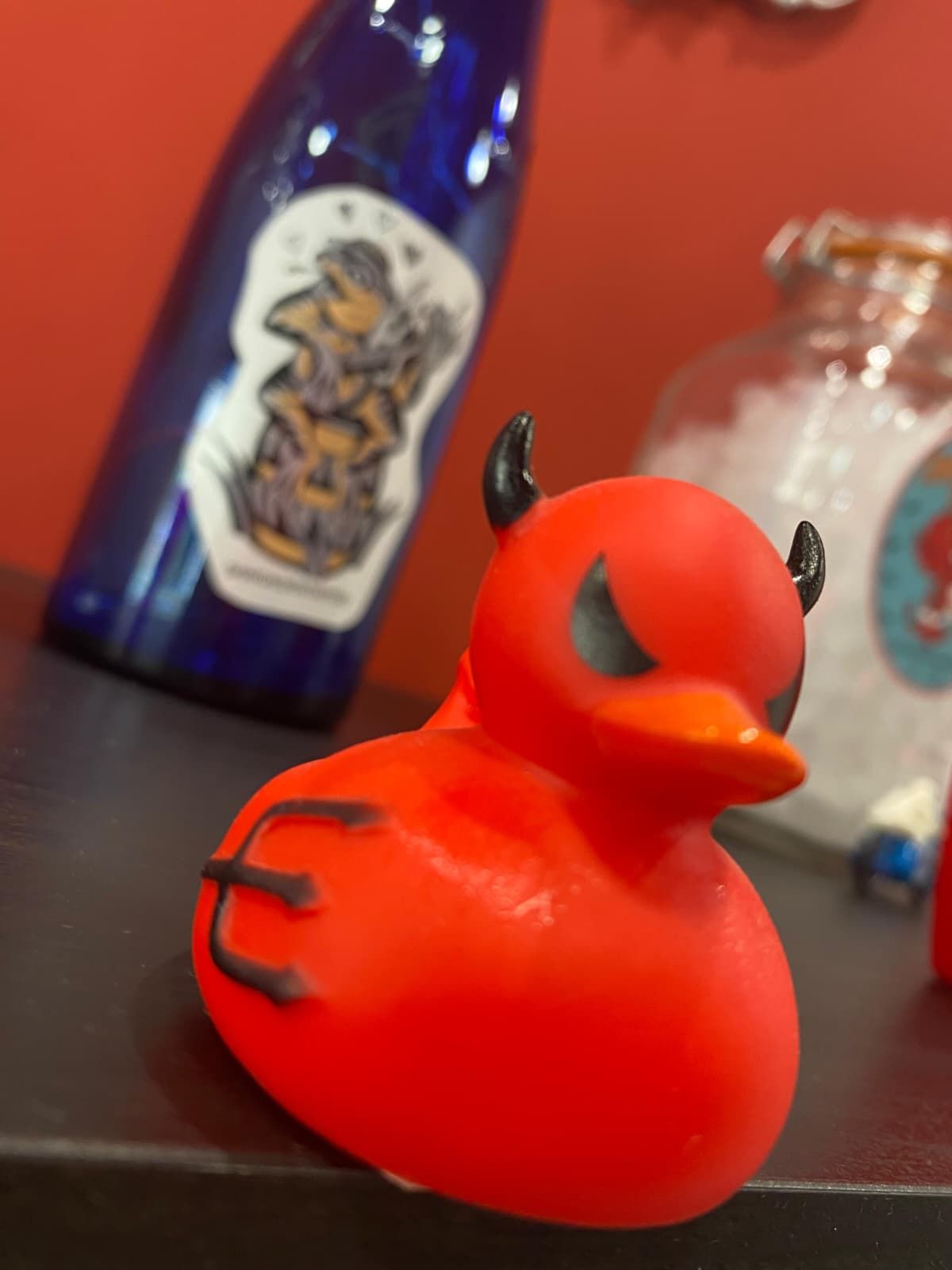 Pato diablo rojo con cuernos y cola negros, en un estante, con una botella azul en el fondo.
