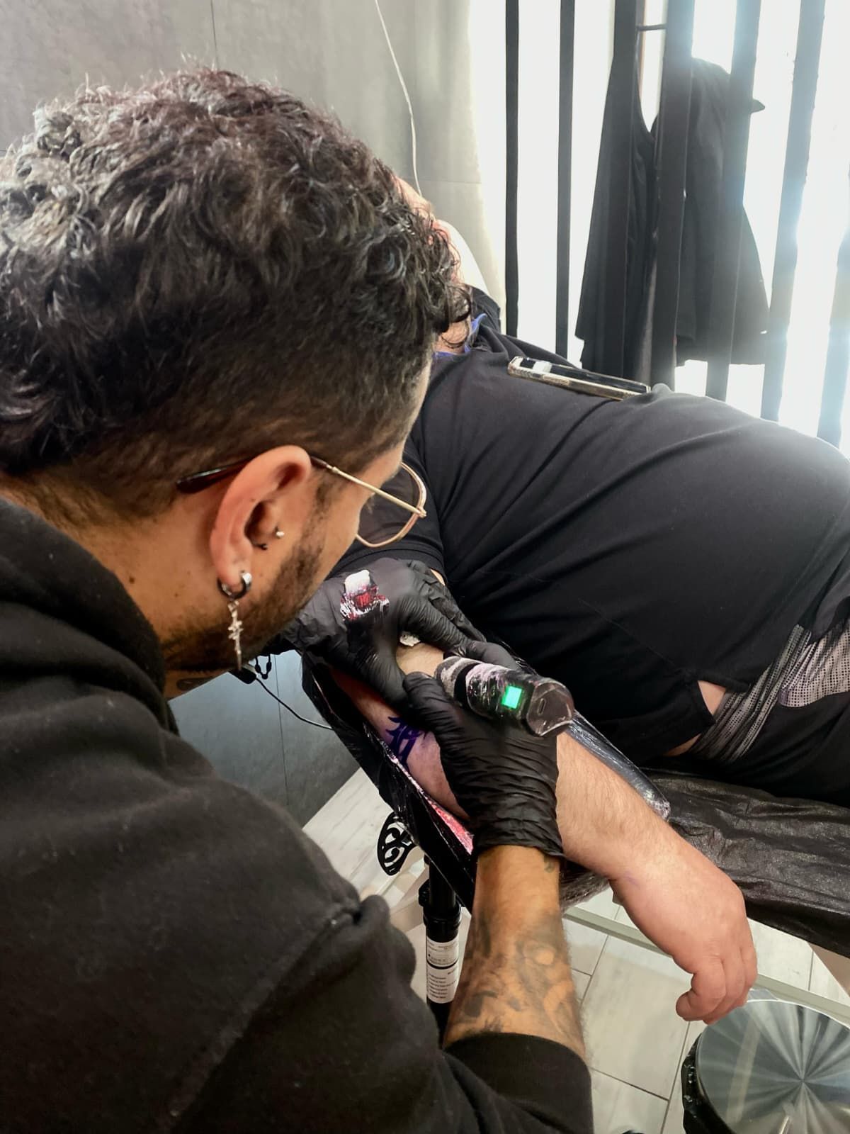 Un artista del tatuaje con gafas y guantes negros entinta el brazo de una persona en un estudio.