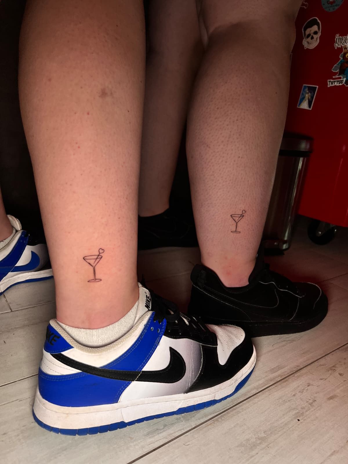 Las piernas de dos personas, cada una con un pequeño tatuaje de una copa de cóctel en el tobillo, calzando zapatillas Nike.