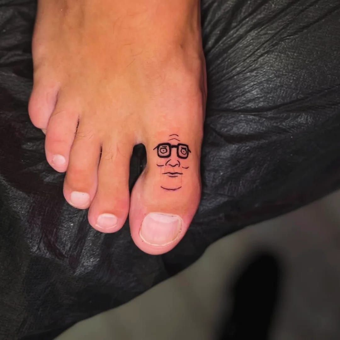 Tatuaje de una cara con gafas en un dedo del pie.