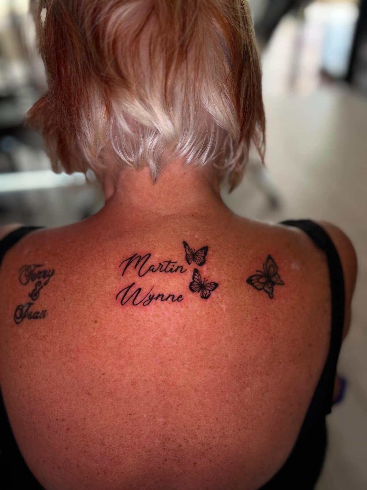Espalda de mujer con tatuajes: nombres y mariposas. Tinta negra sobre piel clara, cabello rubio corto.